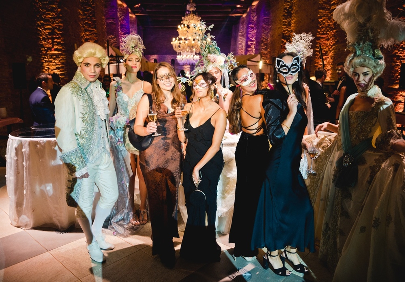 Group in ball gowns and venitian masks holding drinks
